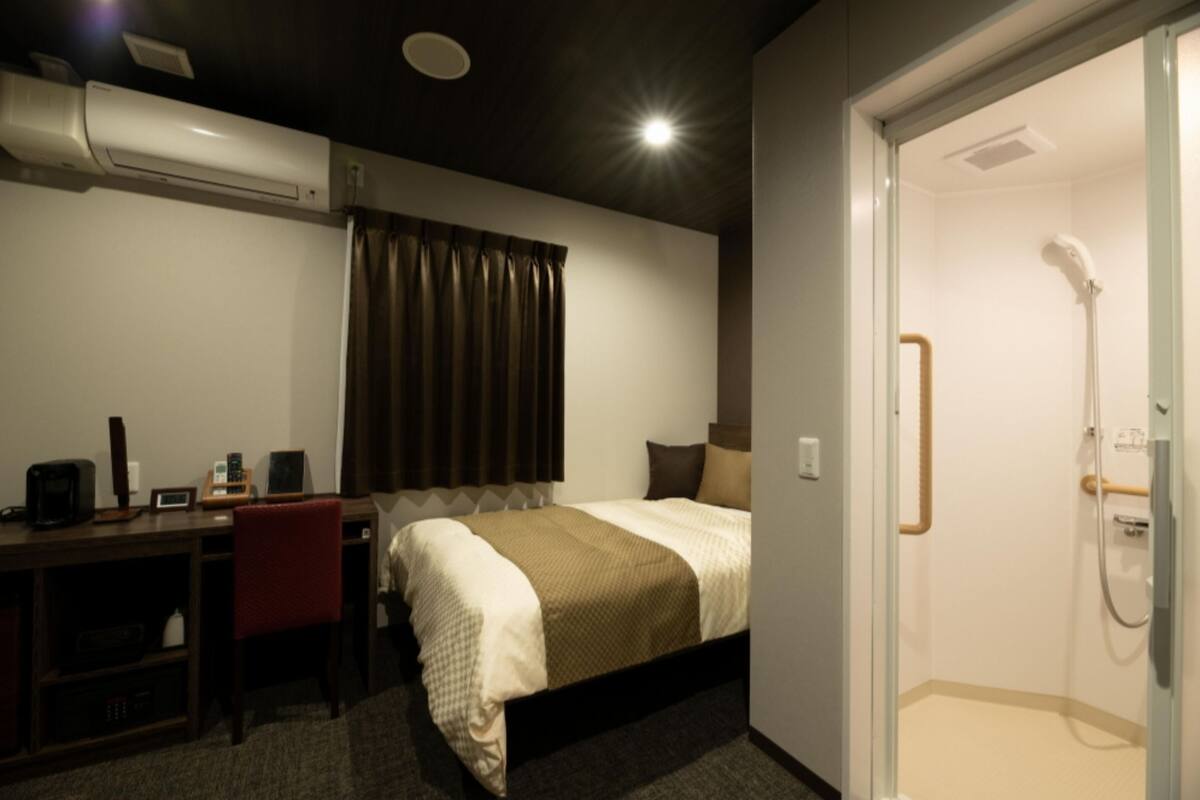 Habitación de hotel con cama, escritorio, silla y zona de ducha.