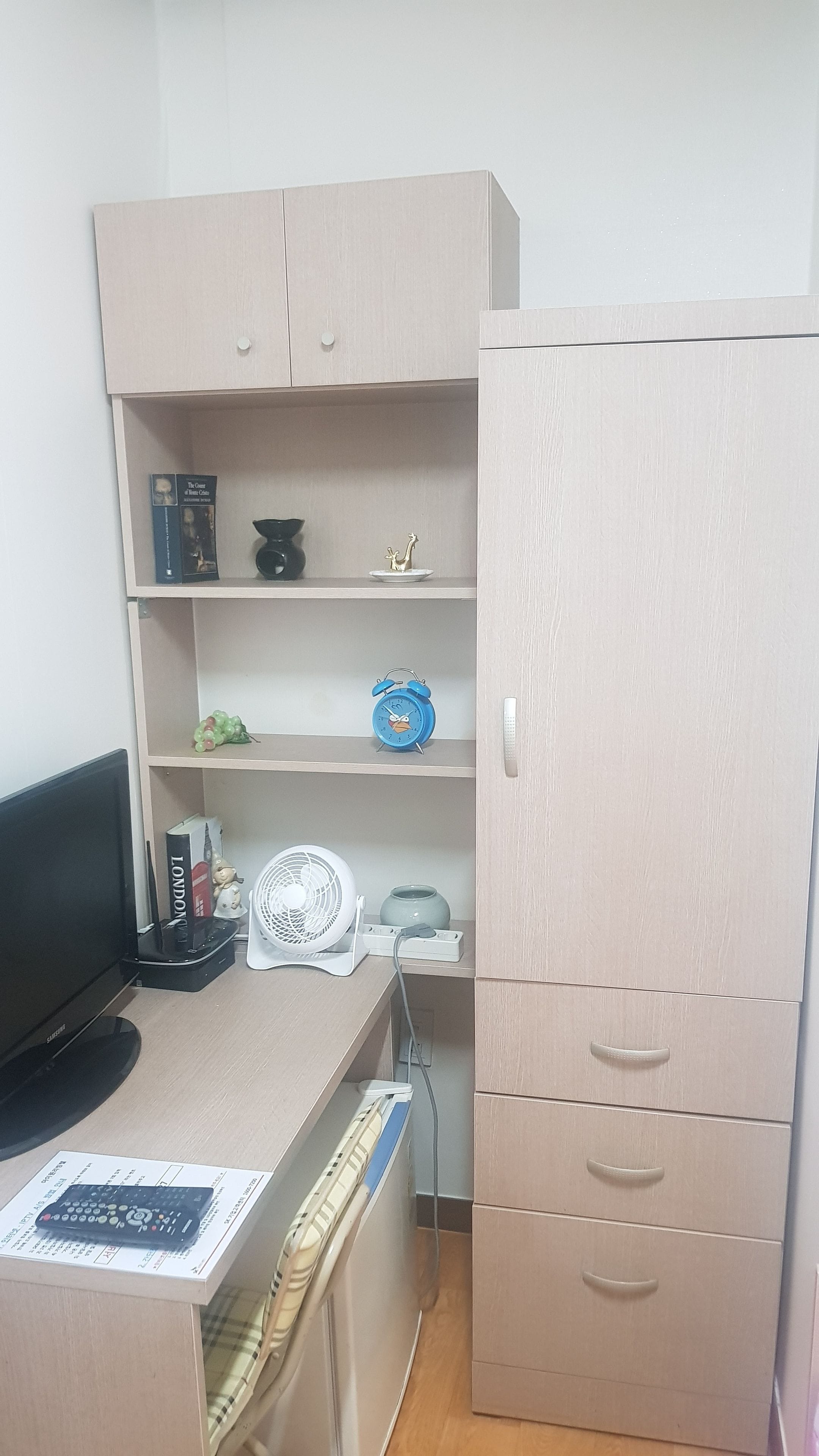 Una habitación con escritorio, computadora, ventilador, estantería y armario.