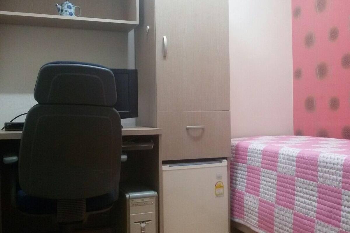 Une petite pièce comprenant un bureau, une chaise et un lit recouvert d’une couette rose.