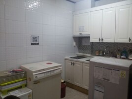 Una kitchenette pequeña con armarios blancos, un microondas y un lavabo. Hay un código QR en la pared y un pequeño cubo de basura.