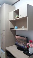 Una habitación con un escritorio color beige, un monitor de computadora, un florero con flores y un mueble con cajones.