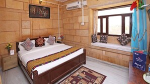 A bedroom with a wooden wall, a bed with patterned pillows, a ceiling fan, an air conditioning unit, a window with curtains, and a bedside table with a vase of flowers.
