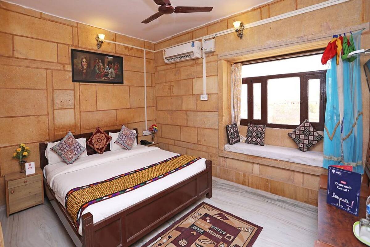 A bedroom with a wooden wall, a bed with patterned pillows, a ceiling fan, an air conditioning unit, a window with curtains, and a bedside table with a vase of flowers.
