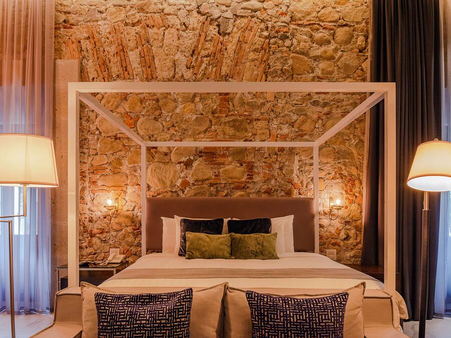 Un lit à baldaquin avec un mur en pierre en arrière-plan, deux lampes de chevet et des coussins à motifs.