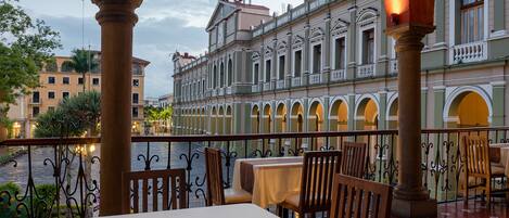 Restaurante con mesas y sillas bajo un área techada con arcos, con vista a un edificio histórico y un río al fondo.