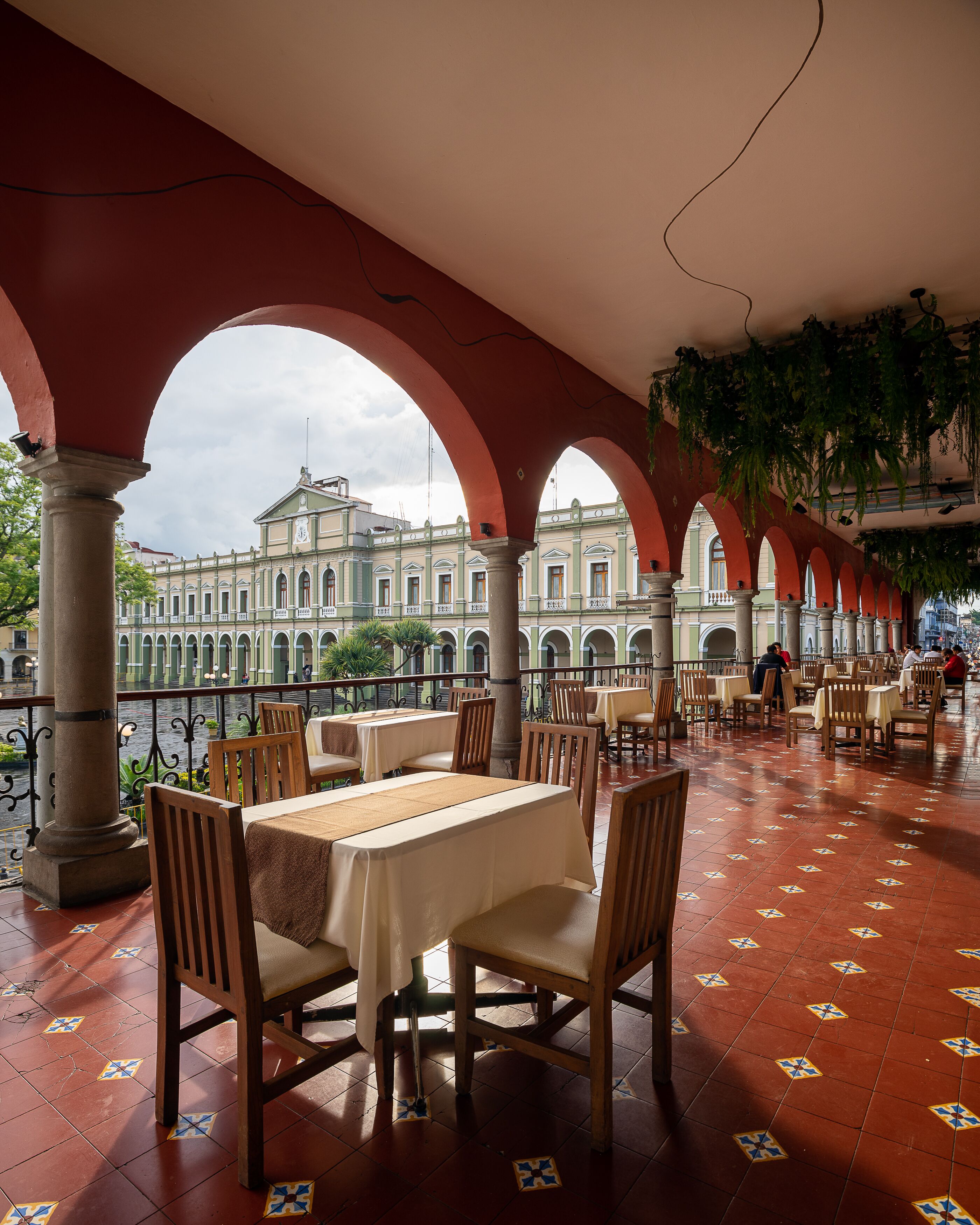 Un área de comedor al aire libre con mesas y sillas, piso de baldosas y vista a un edificio histórico al fondo.