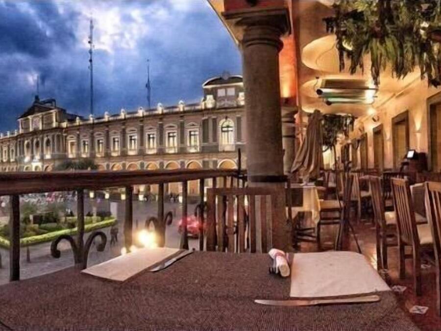 An outdoor dining area with tables and chairs, a view of a historic building, and a railing overlooking a garden.