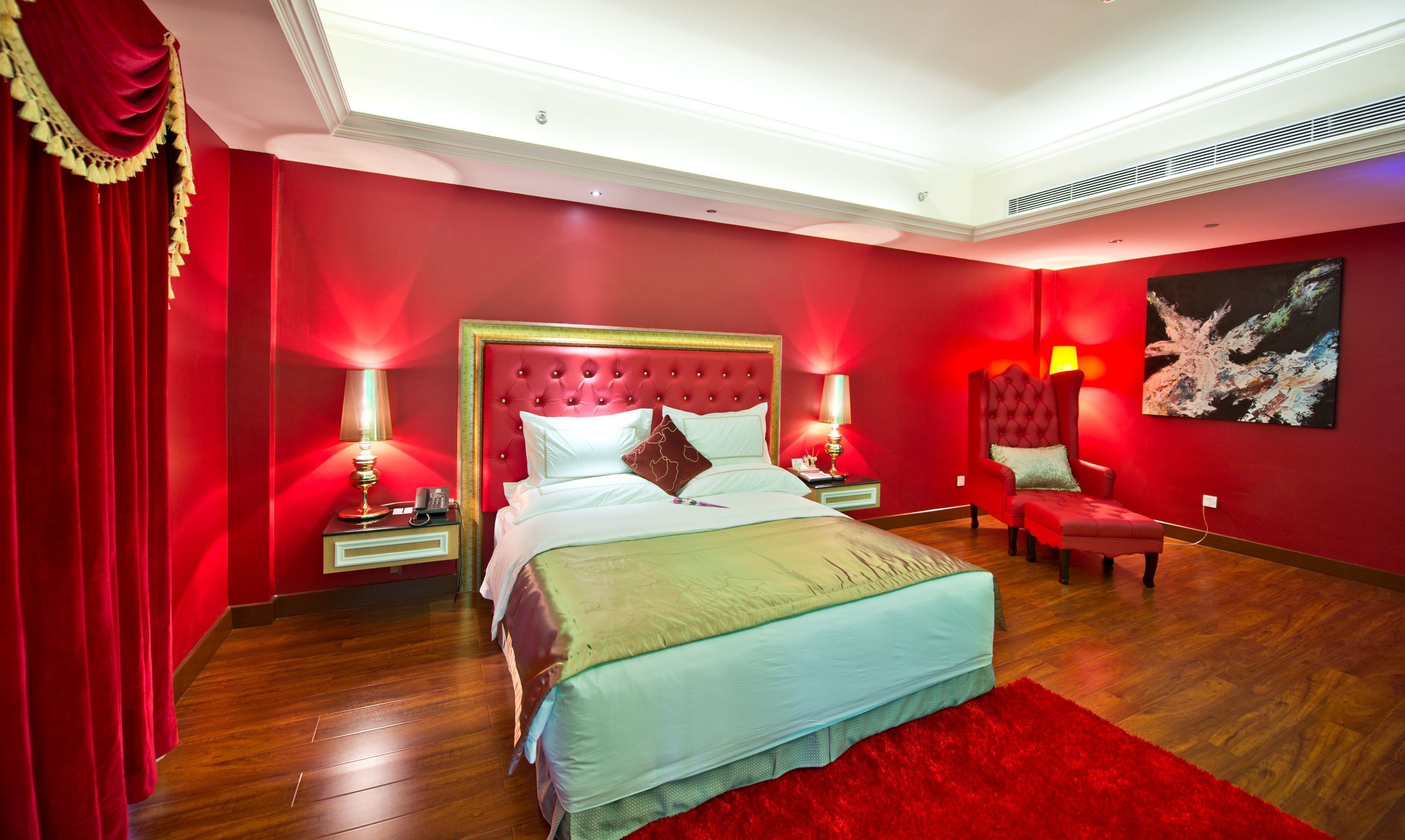 A red-themed bedroom with a large bed, two bedside tables, a red armchair, and a painting on the wall.