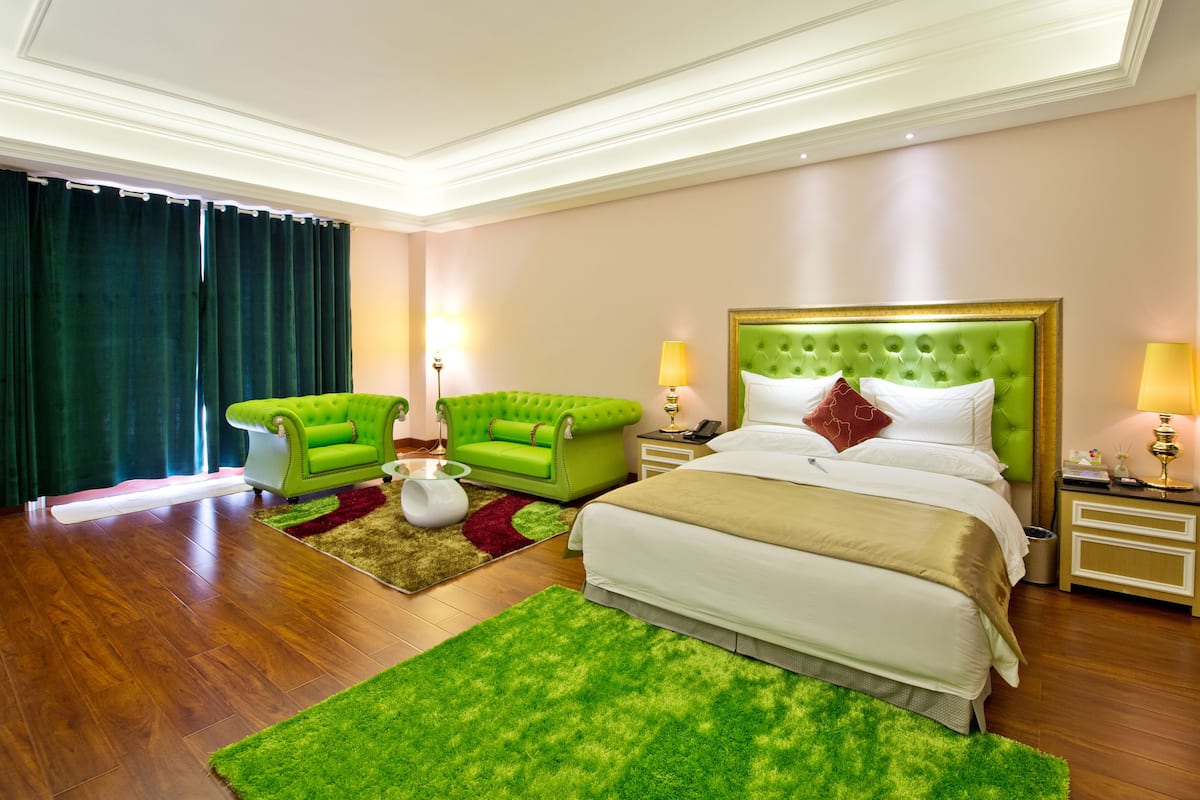 A hotel room with a large bed, two green armchairs, a glass coffee table, and a green area rug.