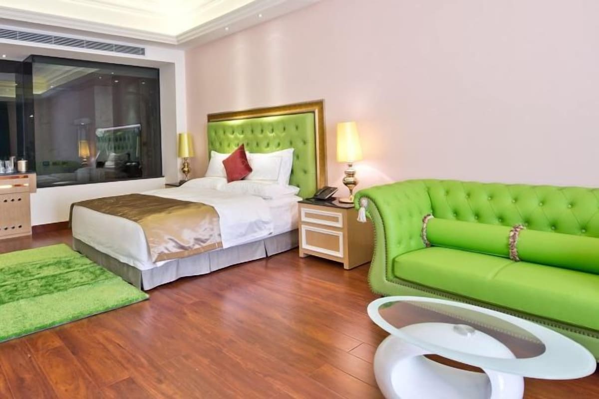 A hotel room with a large bed, a green sofa, a small table, and a window with a view.