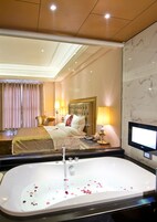 Habitación de hotel con una cama grande, zona de estar con una silla y mesa, televisión y bañera con pétalos de rosa.