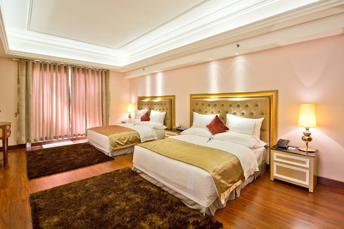 A hotel room with two beds, a wooden floor, a large window with curtains, a bedside table with a lamp, and a small table with a vase.