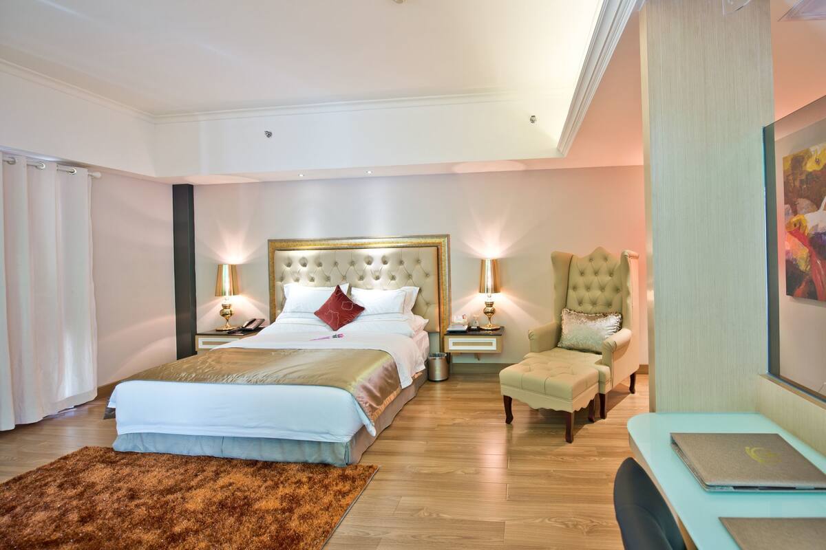 A spacious bedroom with a large bed, a sitting area with a chair, a desk with a laptop, and a painting on the wall.