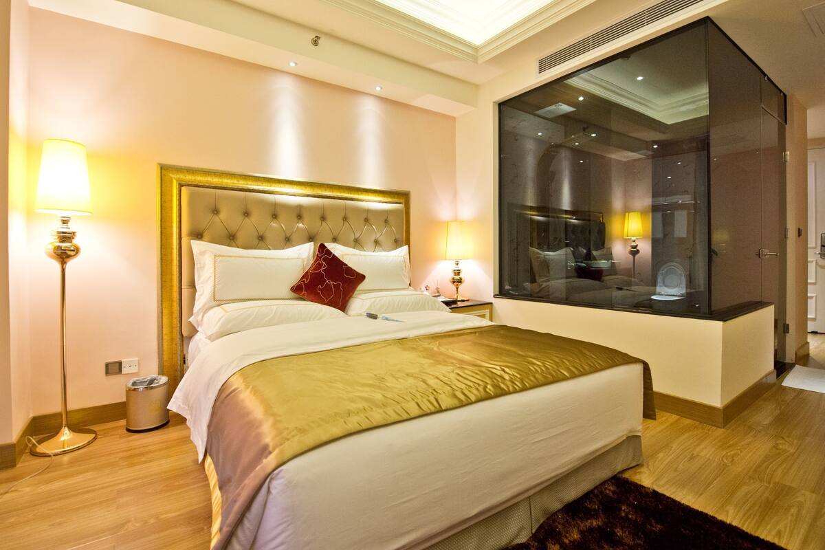 A hotel room with a large bed, two bedside lamps, a mirror, and a view of the city through a large window.
