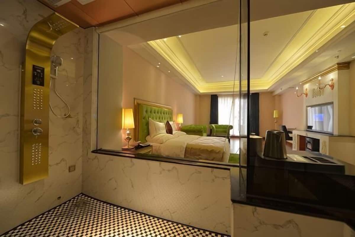 A hotel room with a bed, a mirror, a bathroom with a shower, and a desk with a computer.