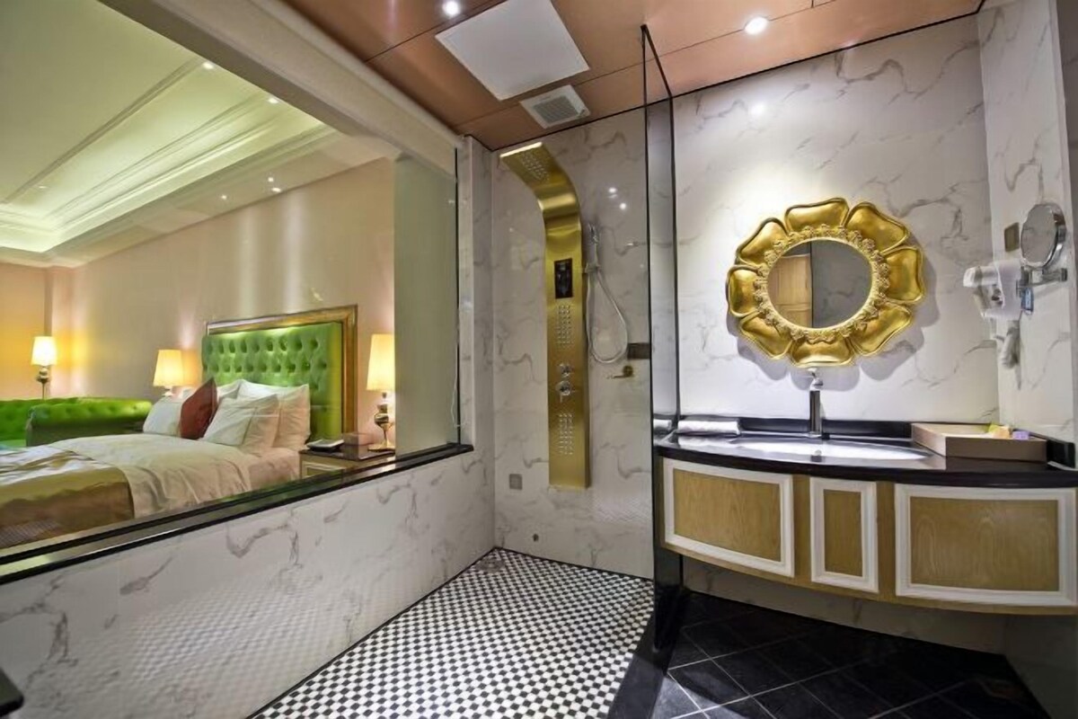 Une chambre d’hôtel avec un mur recouvert de miroirs, une tête de lit recouverte d’un tissu vert, un sol à carreaux noirs et blancs et un mur décoré d’un motif marbré.