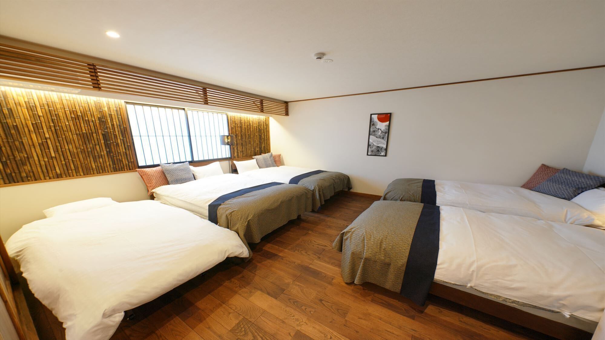A hotel room with three beds, wooden flooring, a wall with vertical blinds, and a ceiling light.