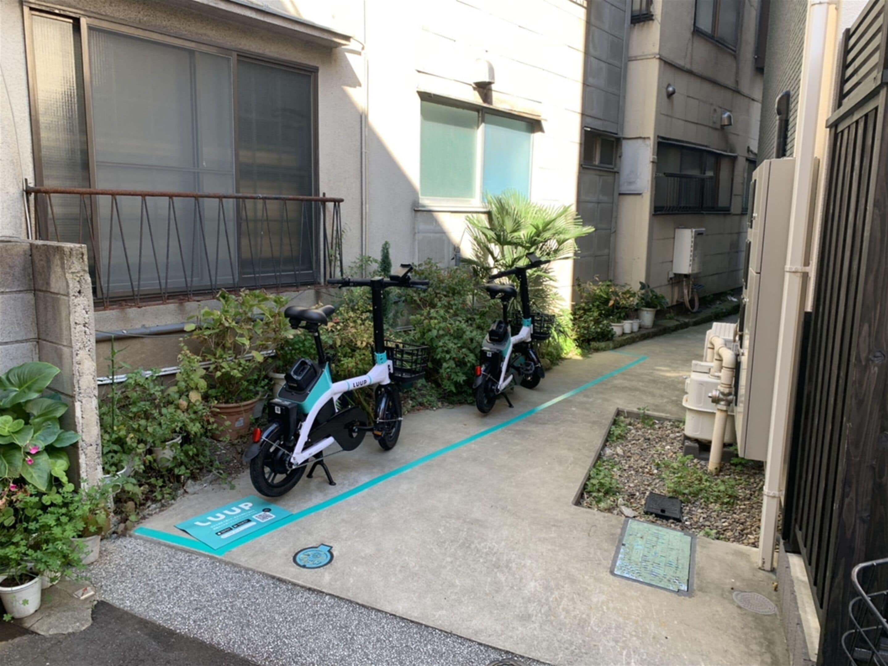 A narrow pathway with two electric scooters parked on the side, surrounded by potted plants and a building with large windows.
