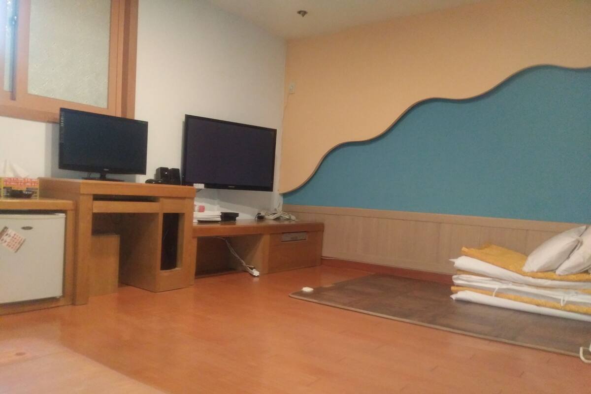 A room with a wooden desk, two televisions, a small refrigerator, and a bed with pillows.