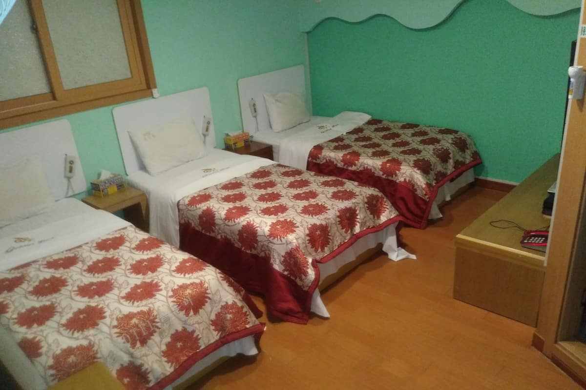 A room with three beds, each with a patterned bedspread, a small bedside table with a lamp, and a wall-mounted shelf with a telephone.
