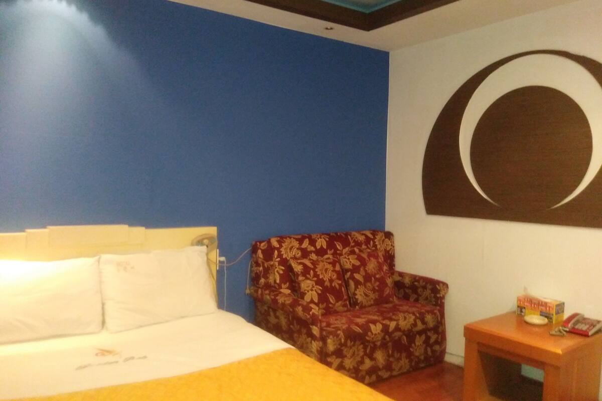 A hotel room with a bed, a floral armchair, a small wooden side table, and a wall with a circular design.