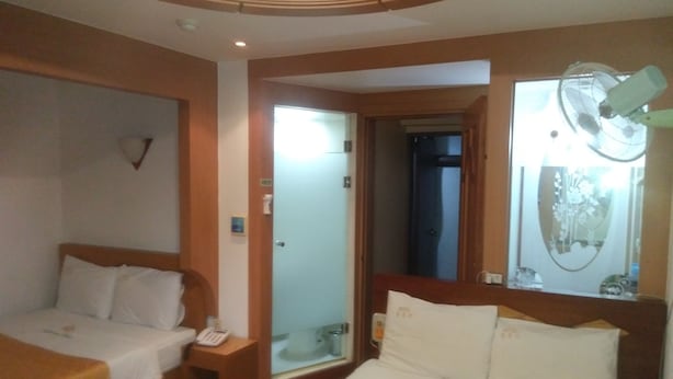 A hotel room with a bed, a bedside table with a phone, a mirror, and a window with a fan. - Golden Bee Motel (Gunsan)