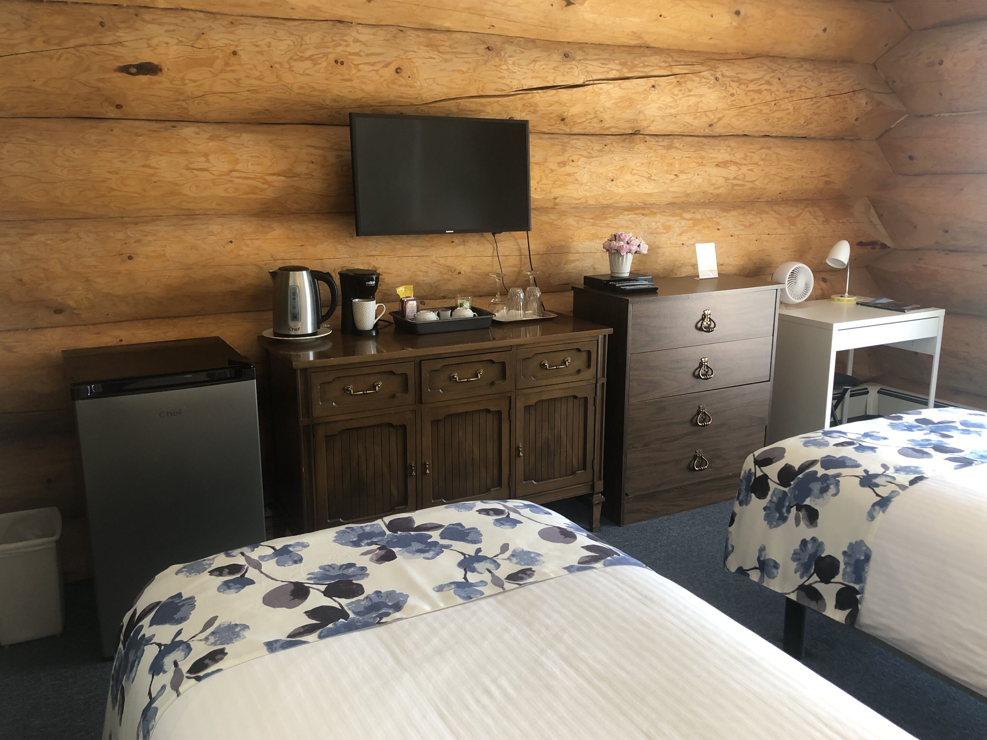 A cozy room with wooden walls, a flat-screen TV mounted above a wooden dresser, a small desk with a fan, and two beds with floral bedding.