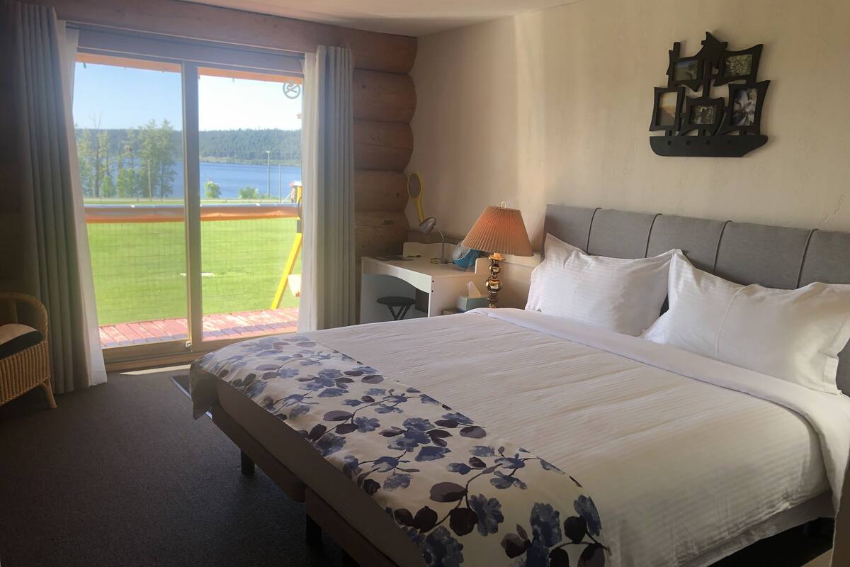 Quarto de hotel com uma cama grande, uma escrivaninha, uma cadeira e vista para um campo verde e água lá fora.