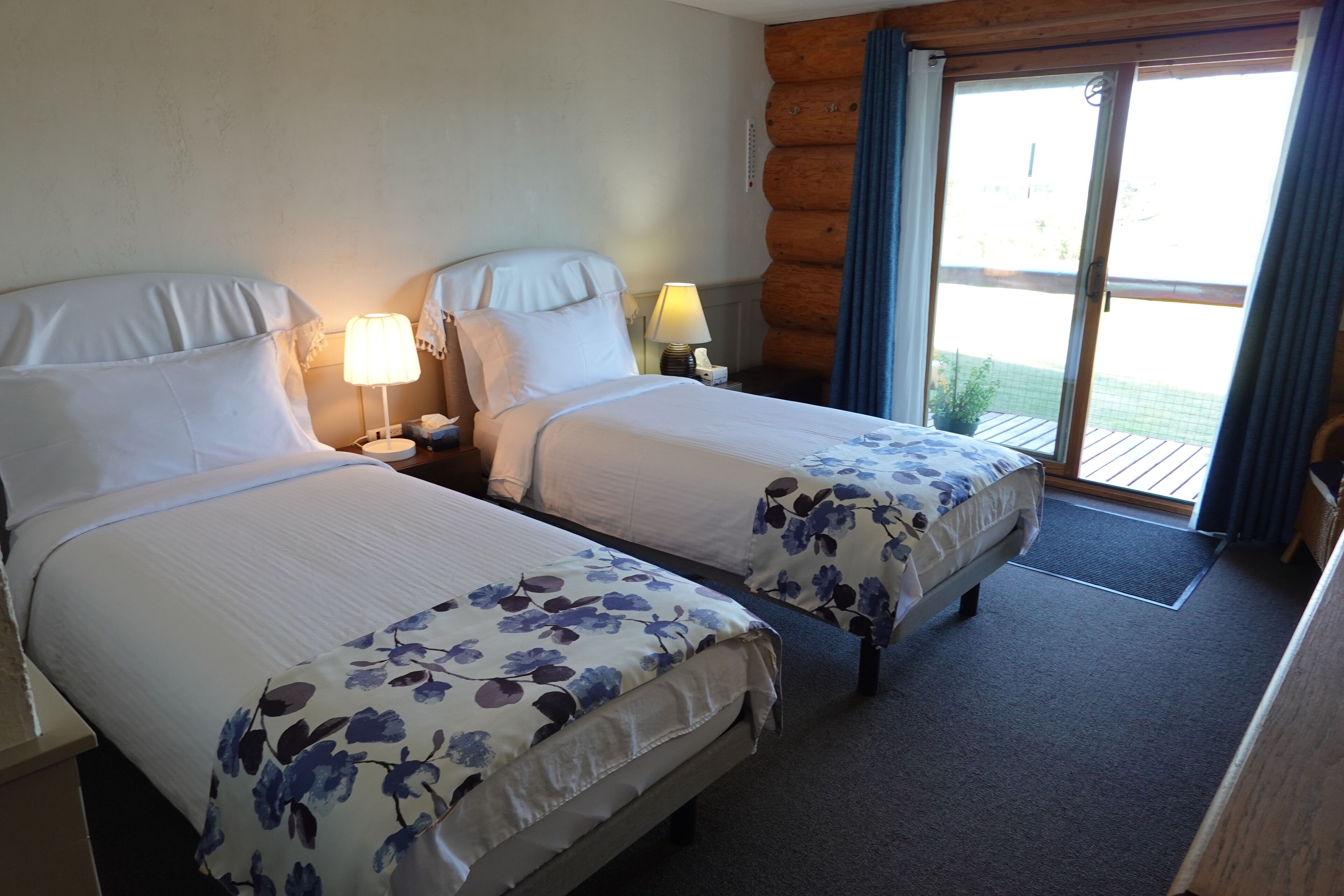 Camera d'albergo con due letti, ciascuno con una coperta a fiori, lampade sui comodini e vista sull'esterno tramite una porta scorrevole in vetro.