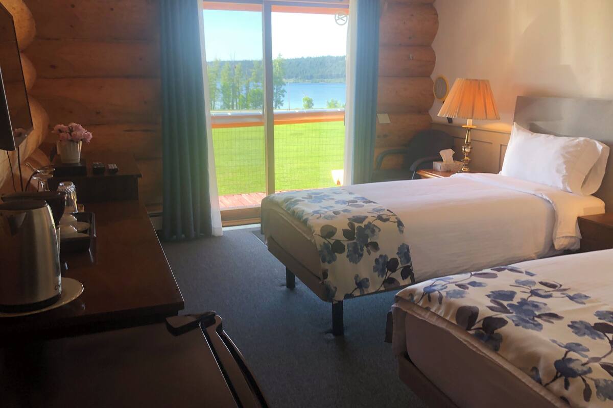 Quarto de hotel com duas camas, uma escrivaninha com abajur, uma cadeira e vista para uma paisagem verde através de uma grande janela.