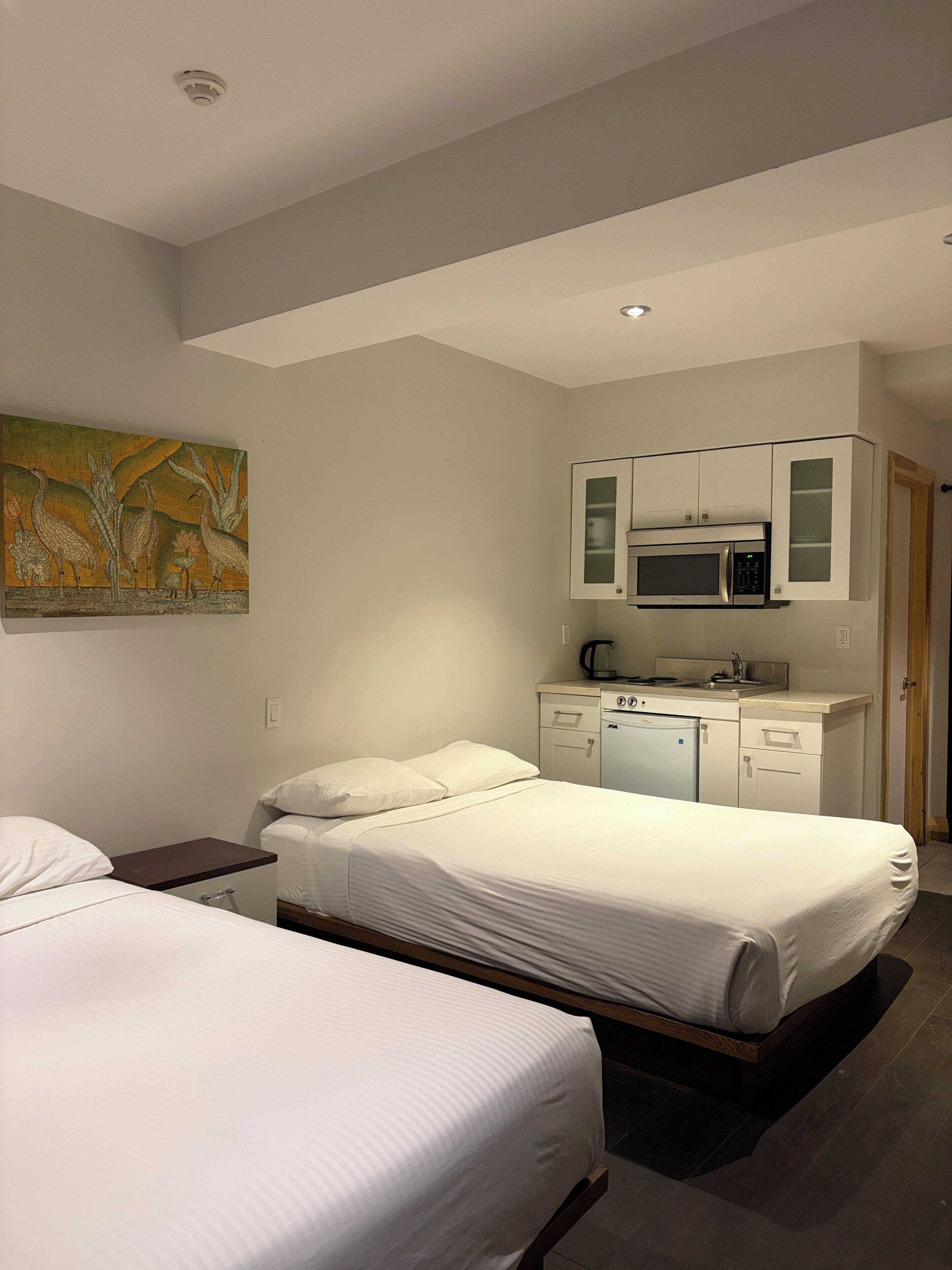 A hotel room with two beds, a kitchenette, and a painting on the wall.