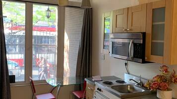 Une cuisine compacte équipée d’un four à micro-ondes, d’une cuisinière et d’un évier, de placards en bois, d’une table à manger en verre avec des chaises et d’une grande fenêtre avec des rideaux.