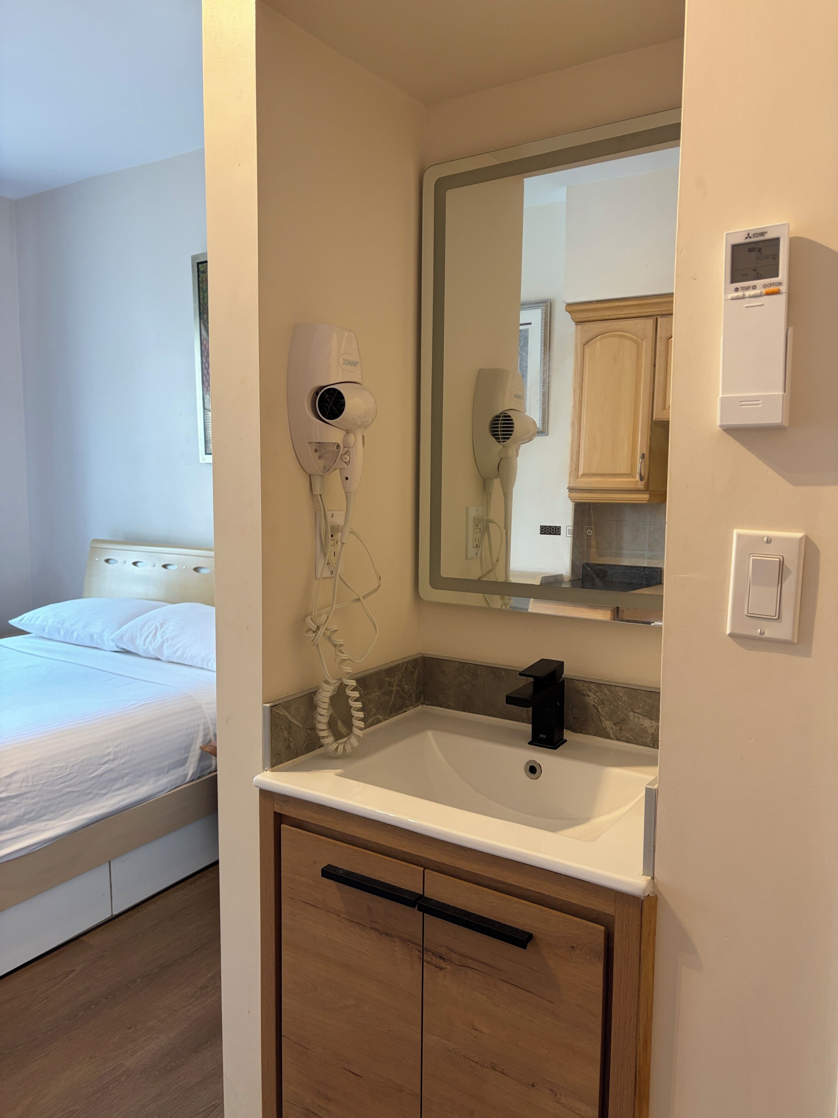 Uma casa de banho de hotel com uma pia, espelho, secador de cabelo e uma cama no quarto adjacente.