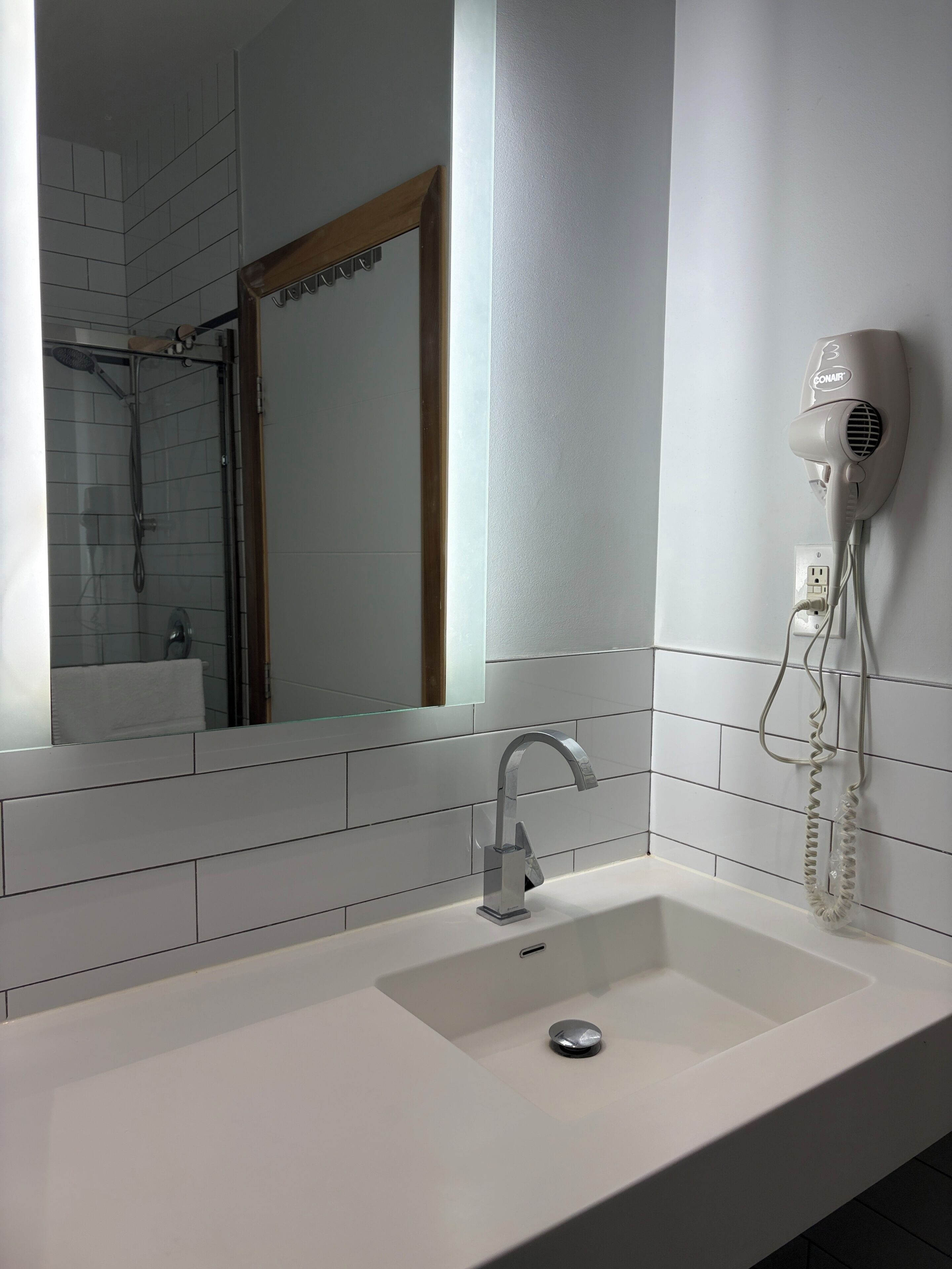 Une salle de bain avec un lavabo blanc, un sèche-cheveux à main fixé au mur et un miroir qui reflète l’espace de la douche.