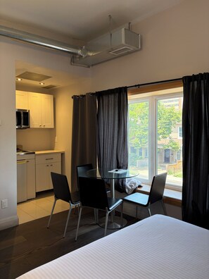 Una habitación de hotel con una cama, una mesita pequeña, dos sillas, un ventanal con cortinas, una kitchenette y un equipo de aire acondicionado.