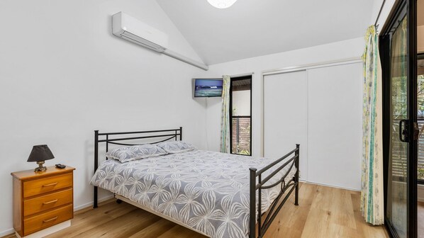 A bedroom with a bed, a TV mounted on the wall, a wooden dresser, and a sliding glass door leading to a balcony. - 43 Satinwood Drive - Rainbow Shores. Pet Friendly and Air Conditioned! (Rainbow Beach)