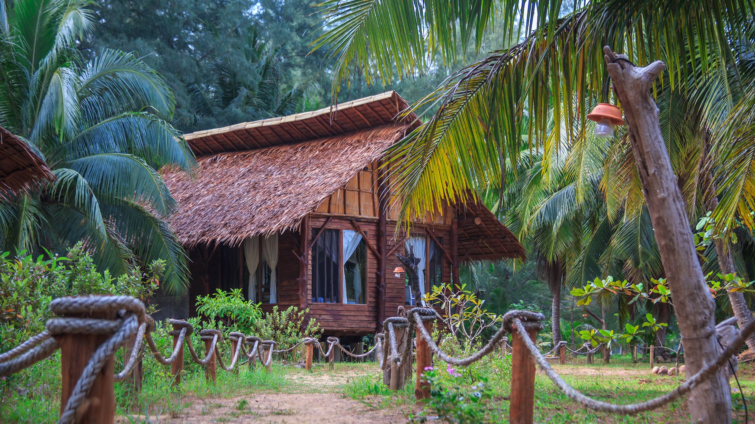 A rustic wooden hut with a thatched roof, surrounded by lush greenery and palm trees.