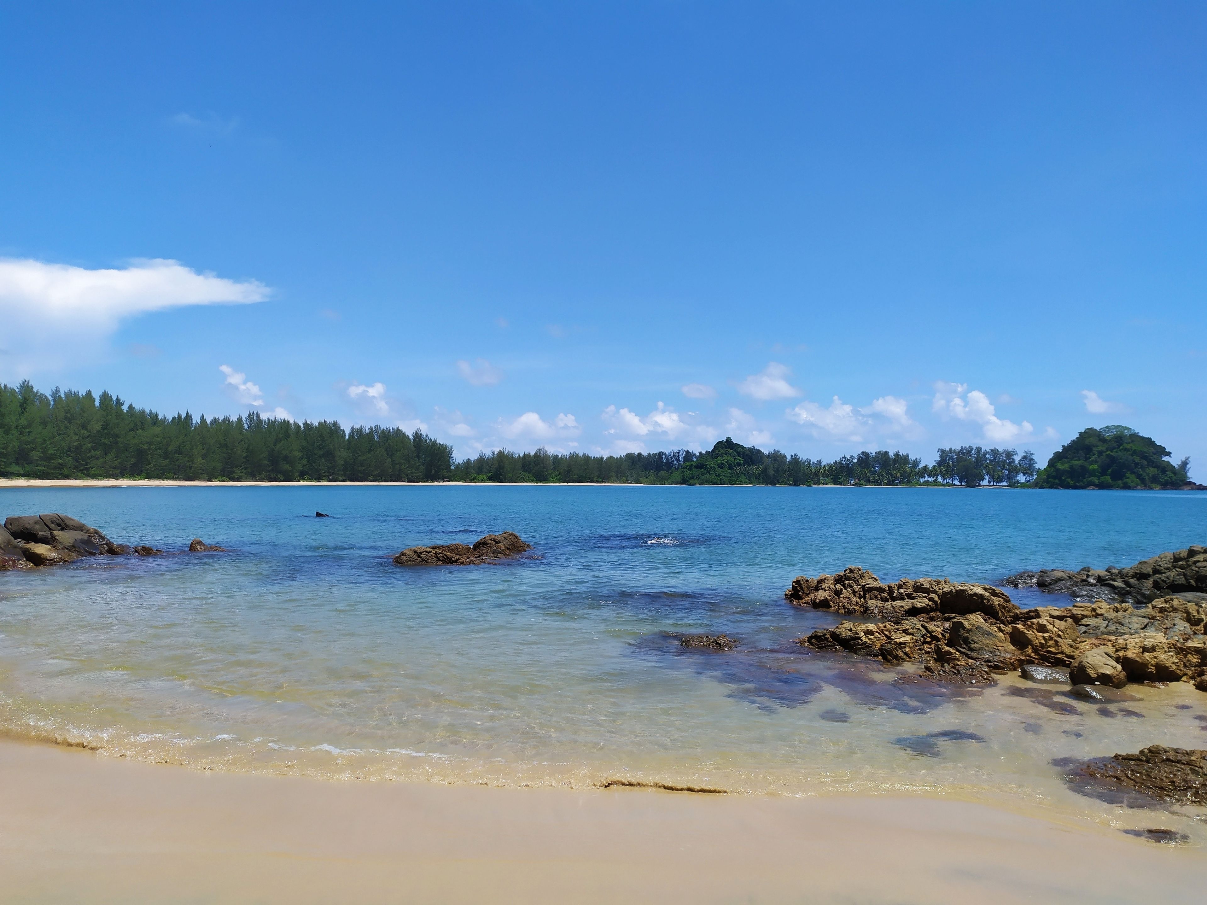 A beach with clear water, rocks, and a forested island in the distance.