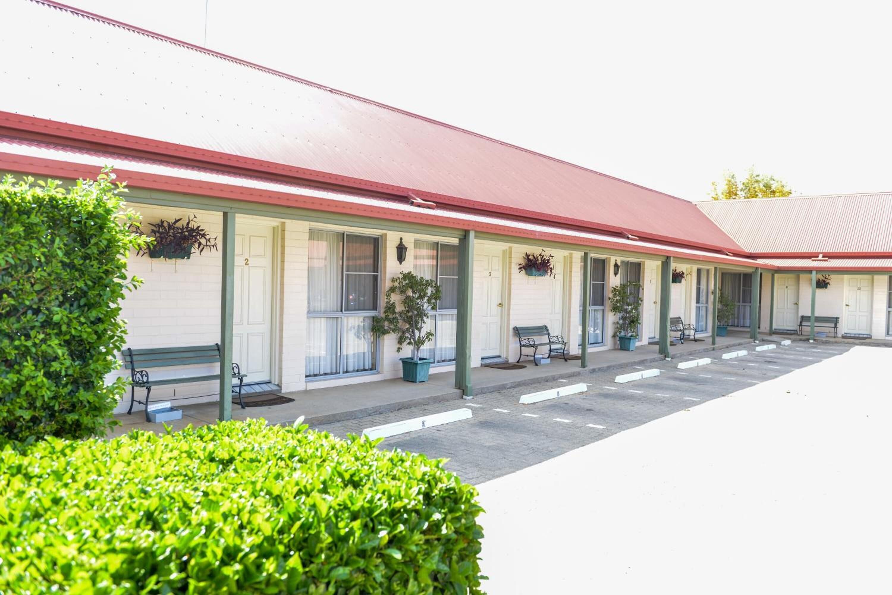 A motel with a row of rooms, each with a window and a door. There are benches and potted plants on the sidewalk.