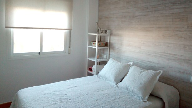A bedroom with a bed, a window with blinds, a shelf with items, and a wooden wall. - Fantastic apartment (El Morche)