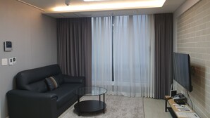 Uma sala de estar moderna com um sofá preto, uma mesa de centro de vidro, uma TV de tela plana e uma mesa lateral de madeira com papéis.