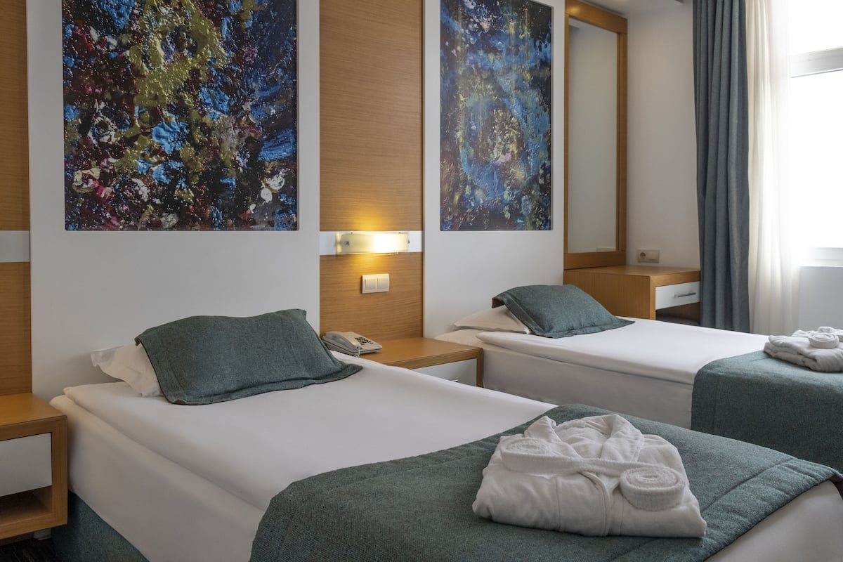 A hotel room with two beds, a wooden headboard, a bedside table with a lamp, and abstract paintings on the wall.