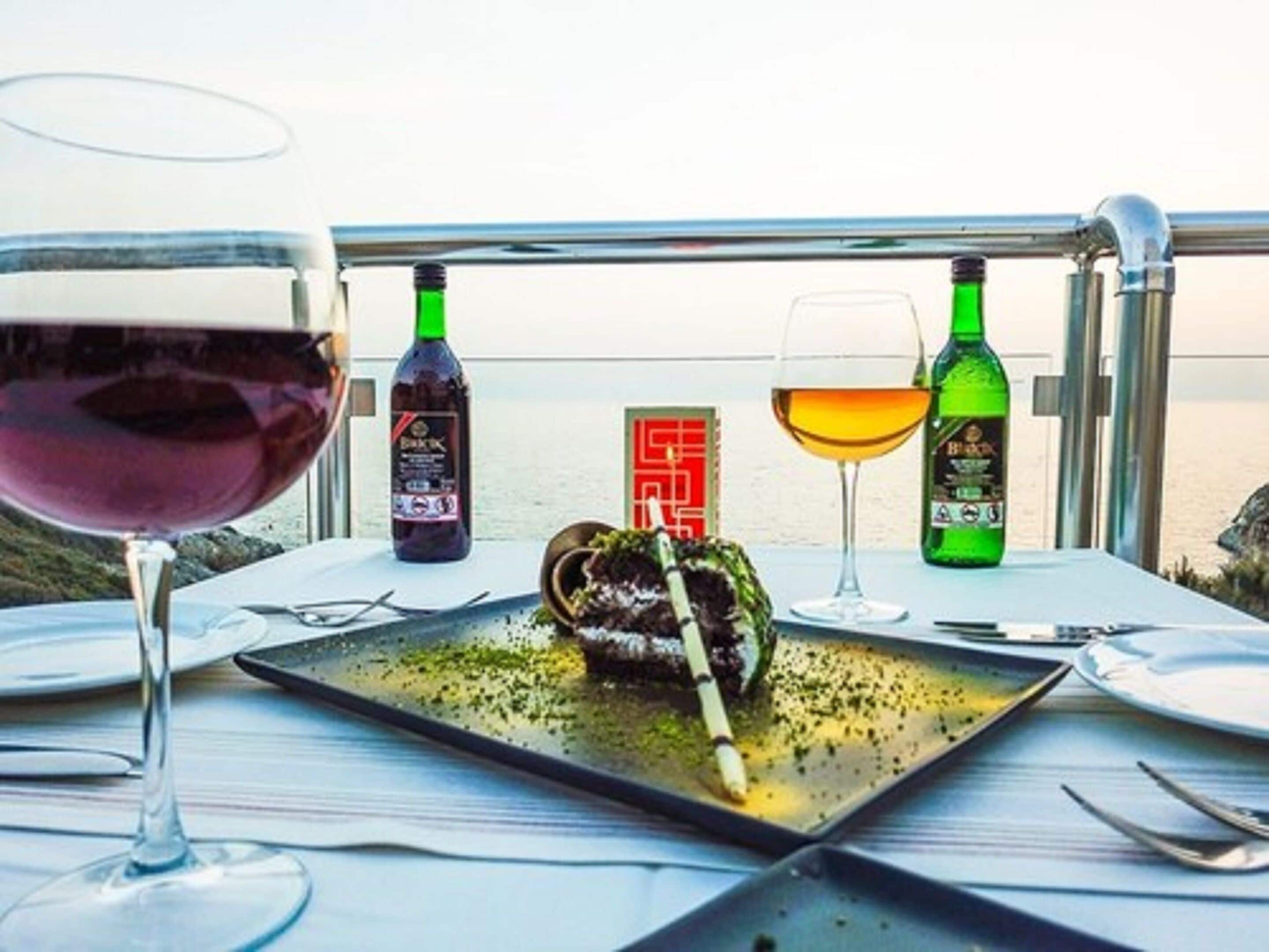 A dining table with a plate of food, two wine glasses, and two bottles of wine.