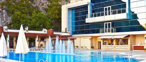 A swimming pool with a water fountain, surrounded by lounge chairs and umbrellas.