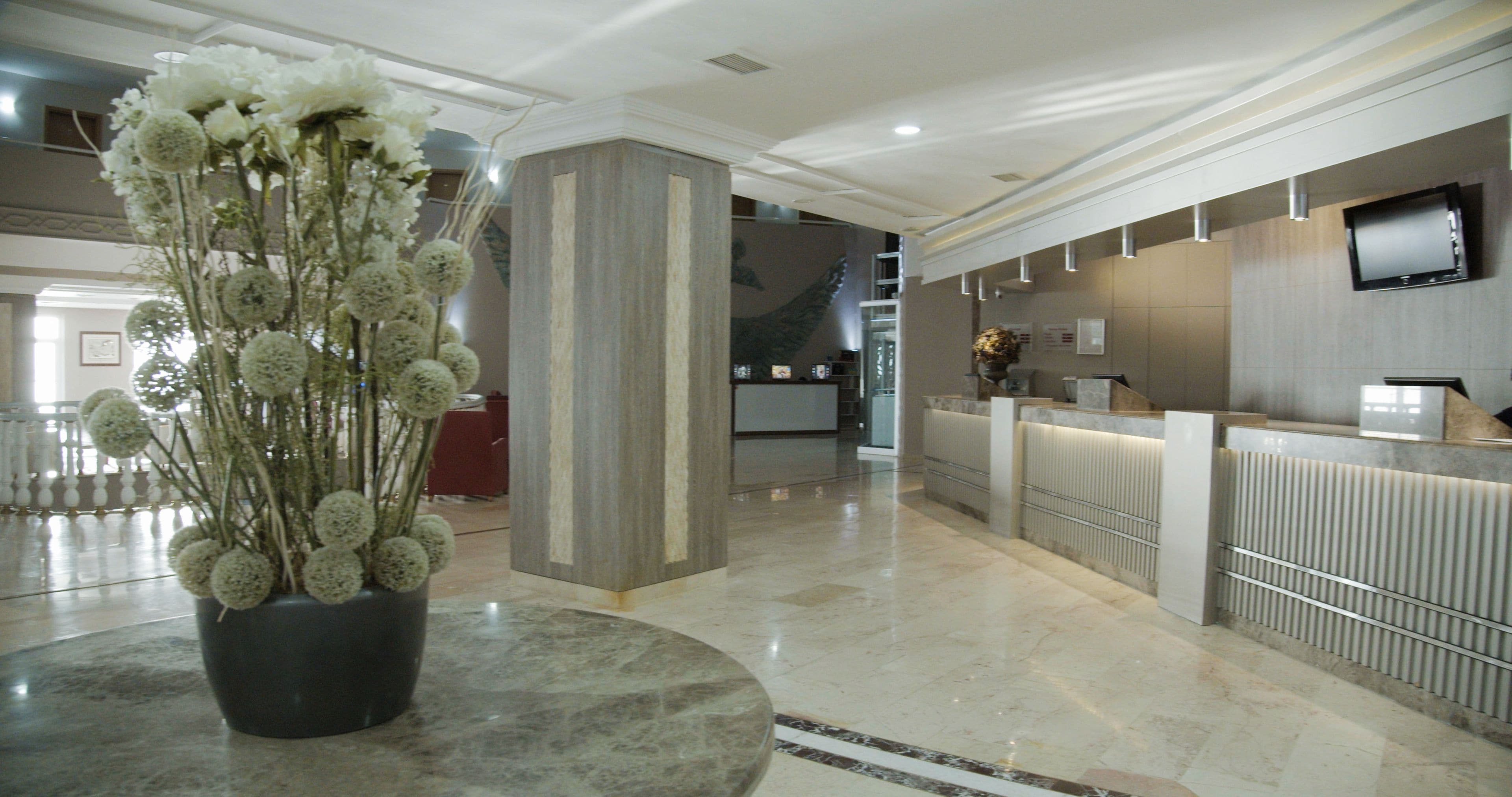 Un hall de hotel con mostrador de recepción, una planta grande en maceta y suelo de mármol.