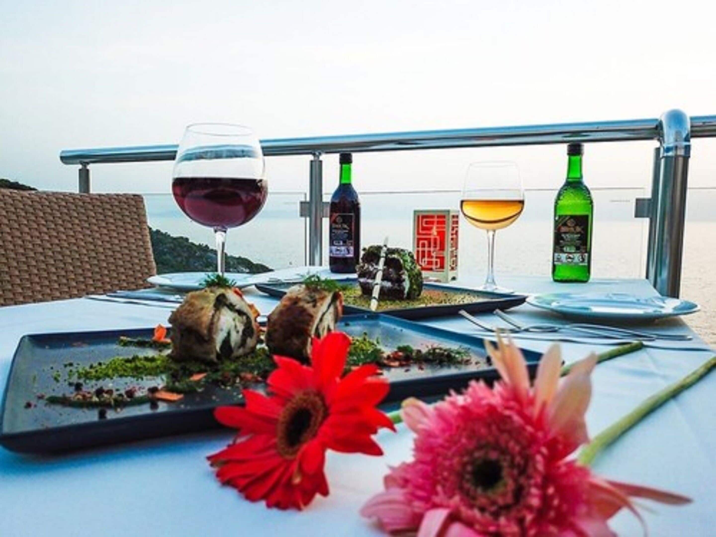 A dining table with a view of the sea, featuring a meal with bread, a red flower, and two wine glasses.
