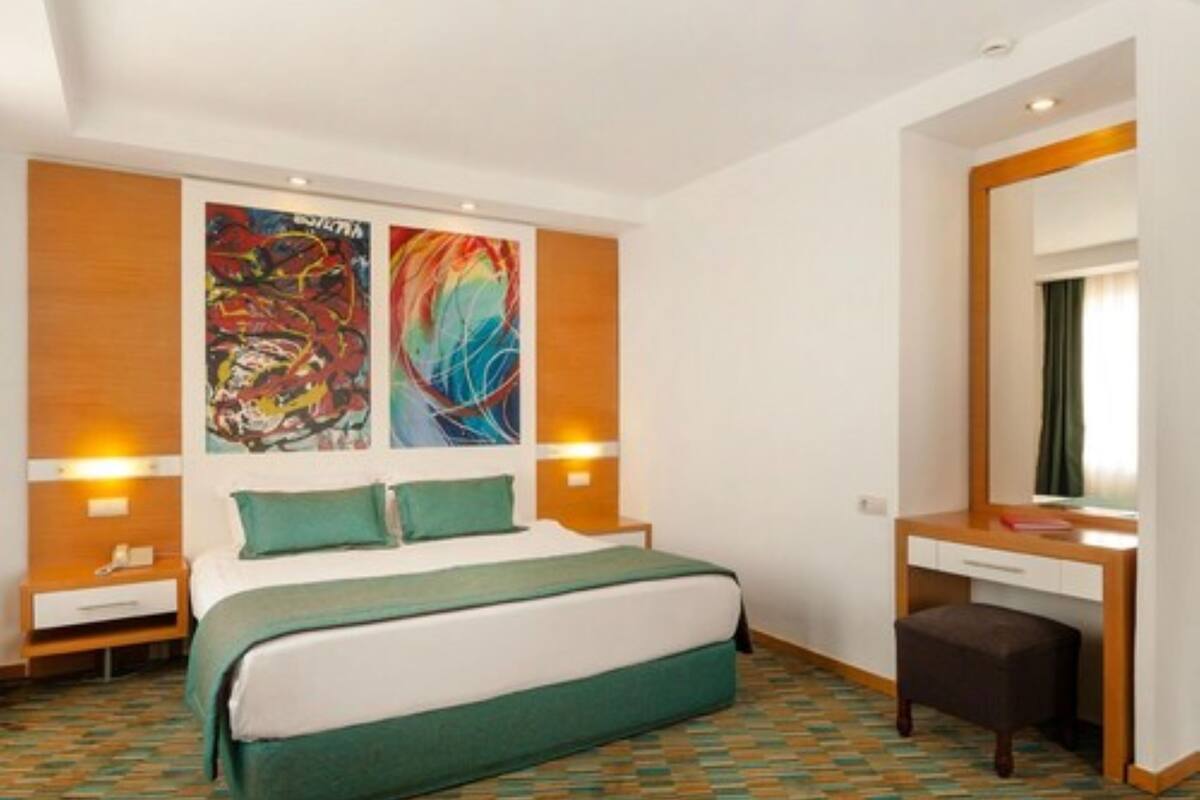 A hotel room with a bed, a desk, and a large mirror. There are two paintings on the wall and a window with curtains.