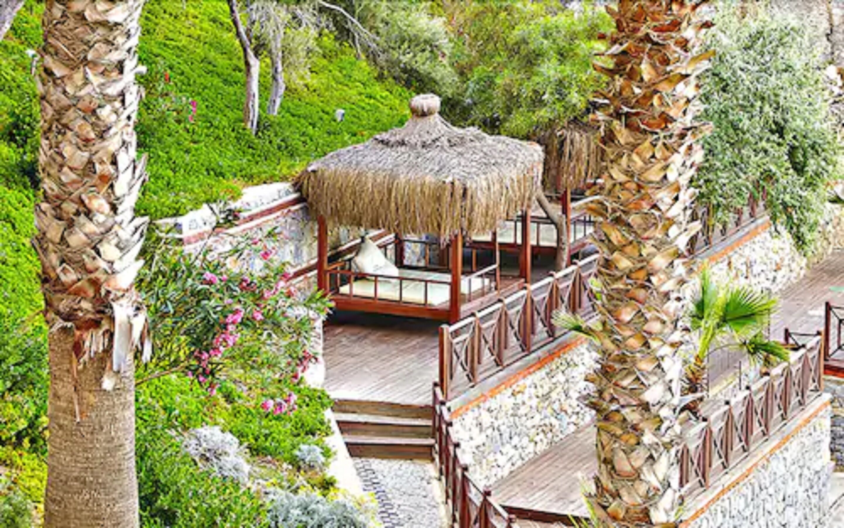 Uma construção com telhado de palha e deck de madeira, rodeada por vegetação exuberante e palmeiras.
