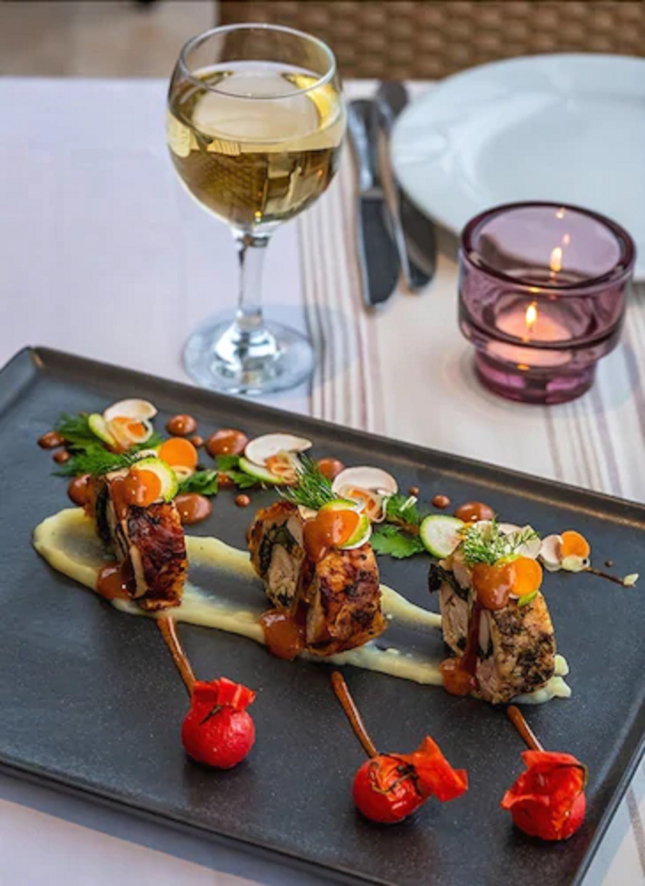 Une assiette avec des brochettes de viande et de légumes, un verre de vin blanc et une bougie sur une table.