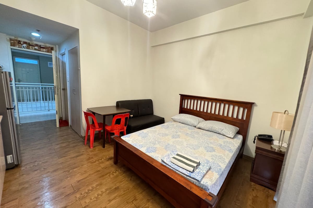 Quarto com uma cama de madeira, uma mesa pequena com duas cadeiras vermelhas, um sofá preto e uma mesa de cabeceira com abajur.